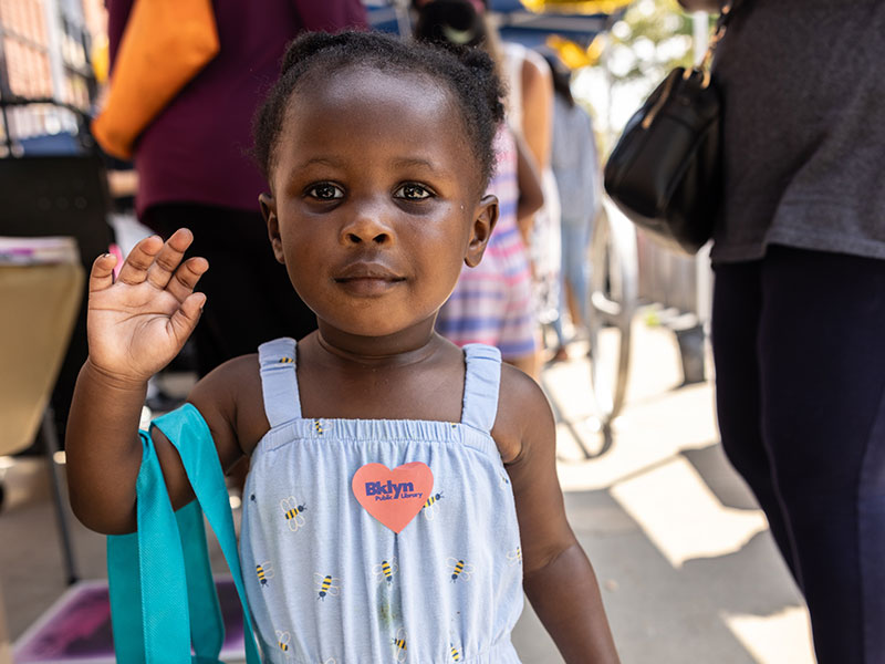 A toddler waves at the camera. She has dark skin and is wearing a bumble bee romper with a BPL sticker stuck to her chest. 