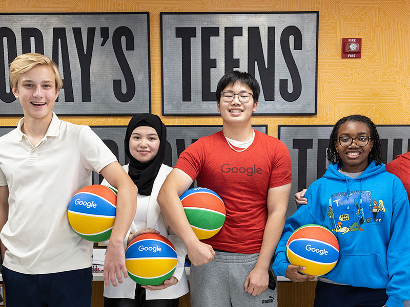 A diverse group of teenagers stand in front of a sign that says Today's Teens, Tomorrow's Techies. They all hold a rainbow basketball with the Google logo on it. 