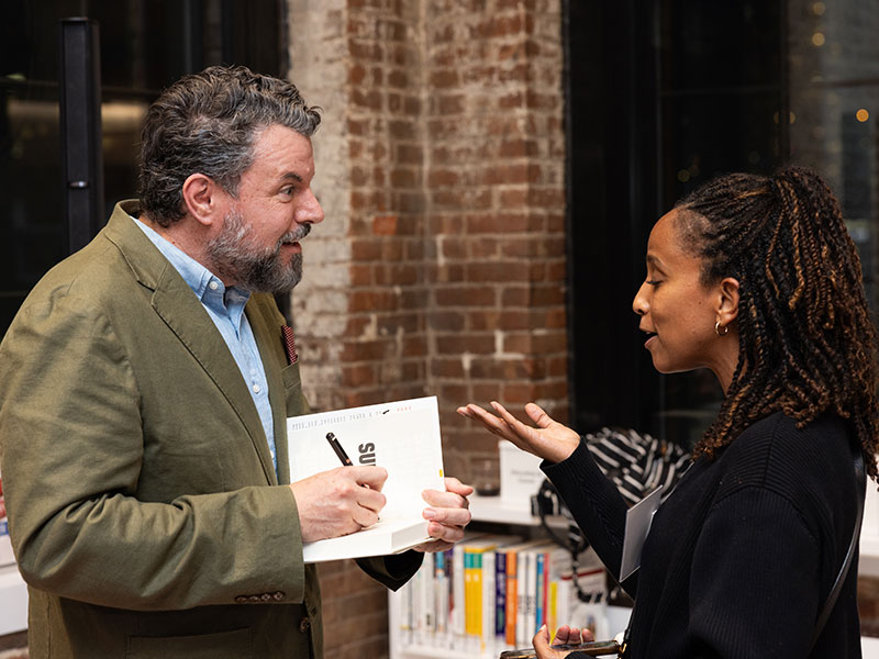 Author Charles Duhigg stands on the left, signing a book for a woman talking to him. Charles is a white man with salt and pepper hair and a beard. The woman has dark skin and wears her in twists. The two people are looking at one another and talking animatedly.
