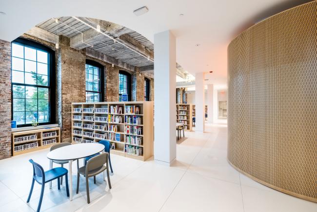 An interior photograph of Adams Street Library. The right side of the photo is a curved wall and on the left are bookshelves next to a pillar. In the lower left hand corner are a table and chairs.