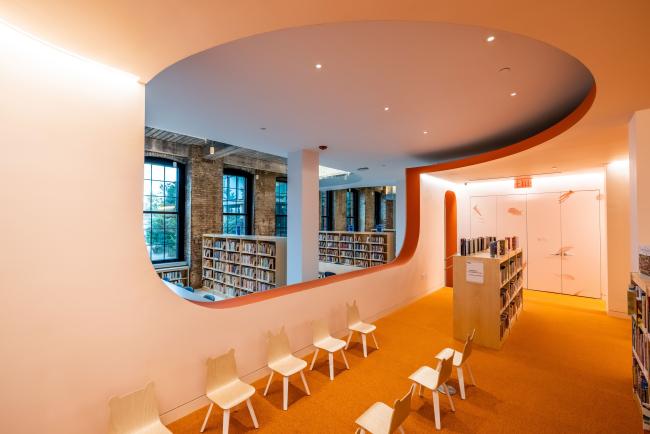 A view of the Adams Street Library children's section looking out into the adult books section through a passthrough window in the center of the room. The carpet is orange and there are two rows of small seats.