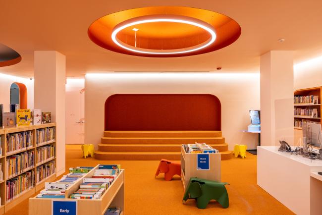 Adams Street Library's children's section: The room is a light orange color with an orange carpet. There are low tables and chairs that look like elephants in the middle of the floor. At the back of the room are low seats.