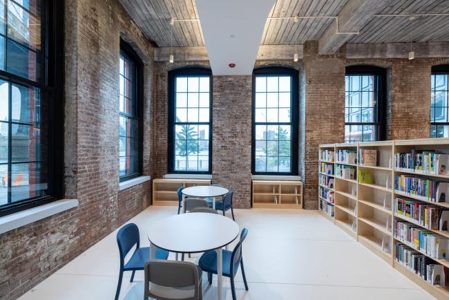 An interior shot of Adams Street Library, facing the windows. In front of the windows are two white tables with four blue chairs at each table.