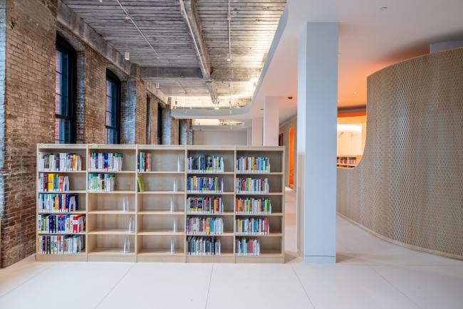 An interior photograph of Adams Street Library, facing the back of the library. In the foreground are bookshelves next to a pillar. In the background you can see the children's section.